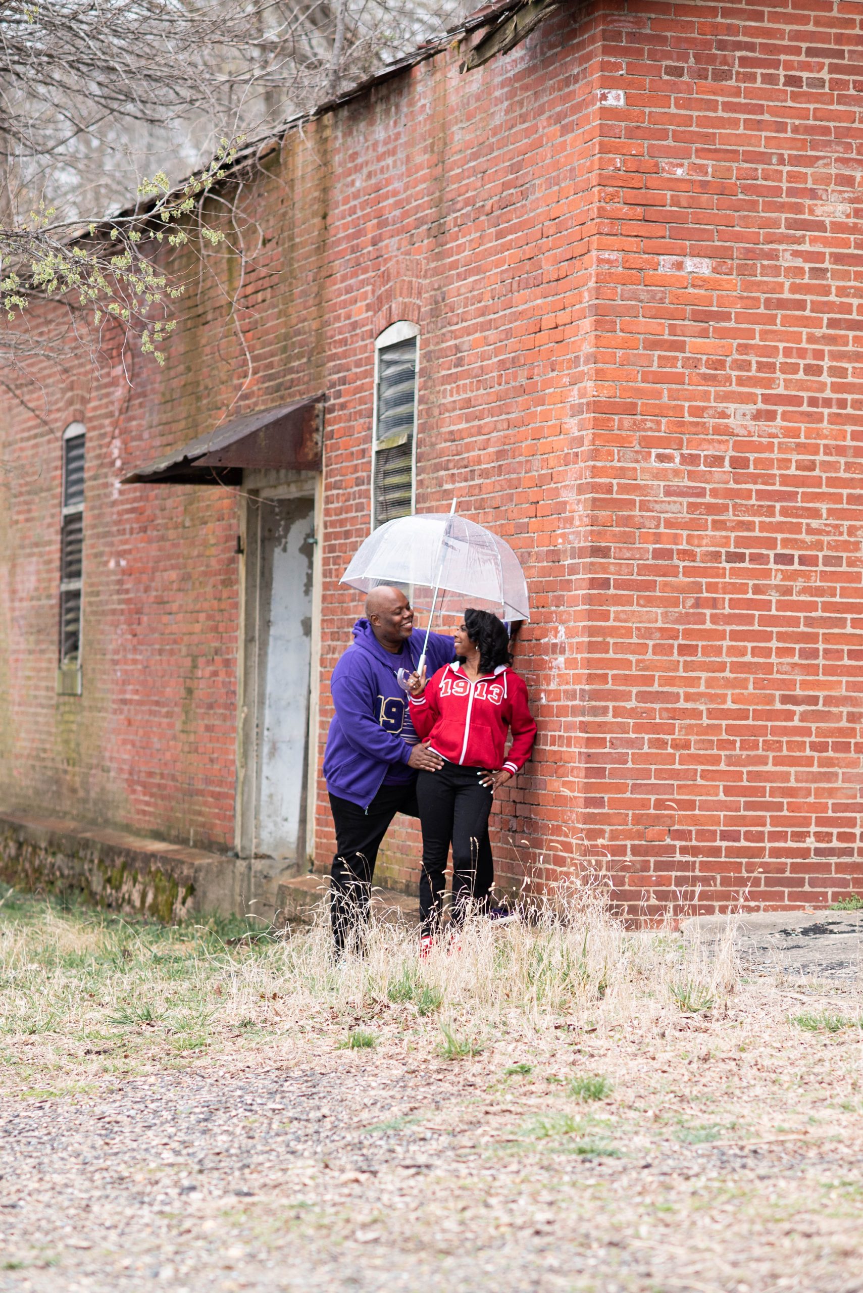 engaged couple poses by brick building under clear umbrella during Maryland engagement photos