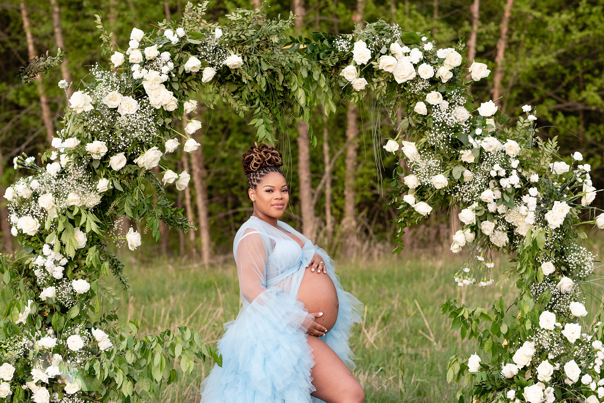 woman in blue dress holds bump under floral arbor