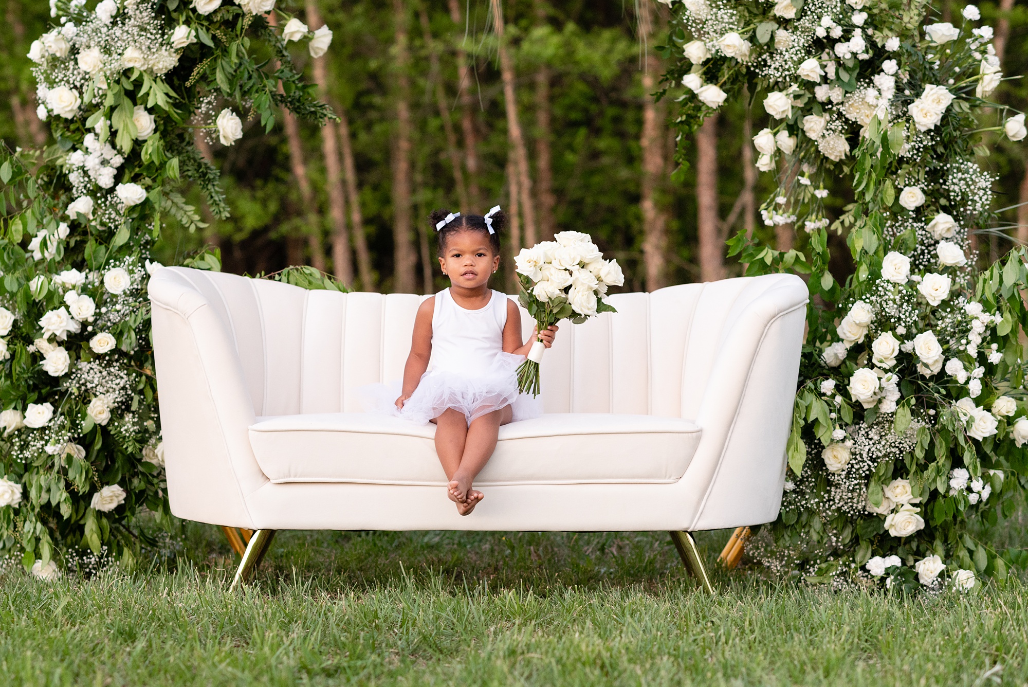 toddler sits on white couch holding bouquet of white roses