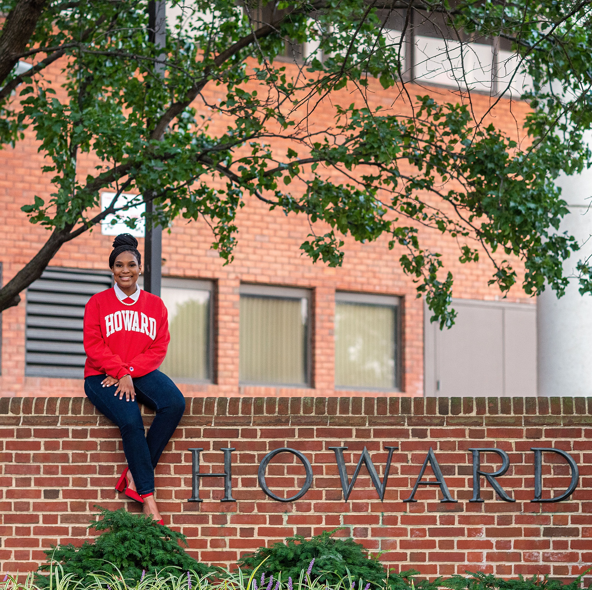 freshman in red sweater sits on brick sign at Howard University